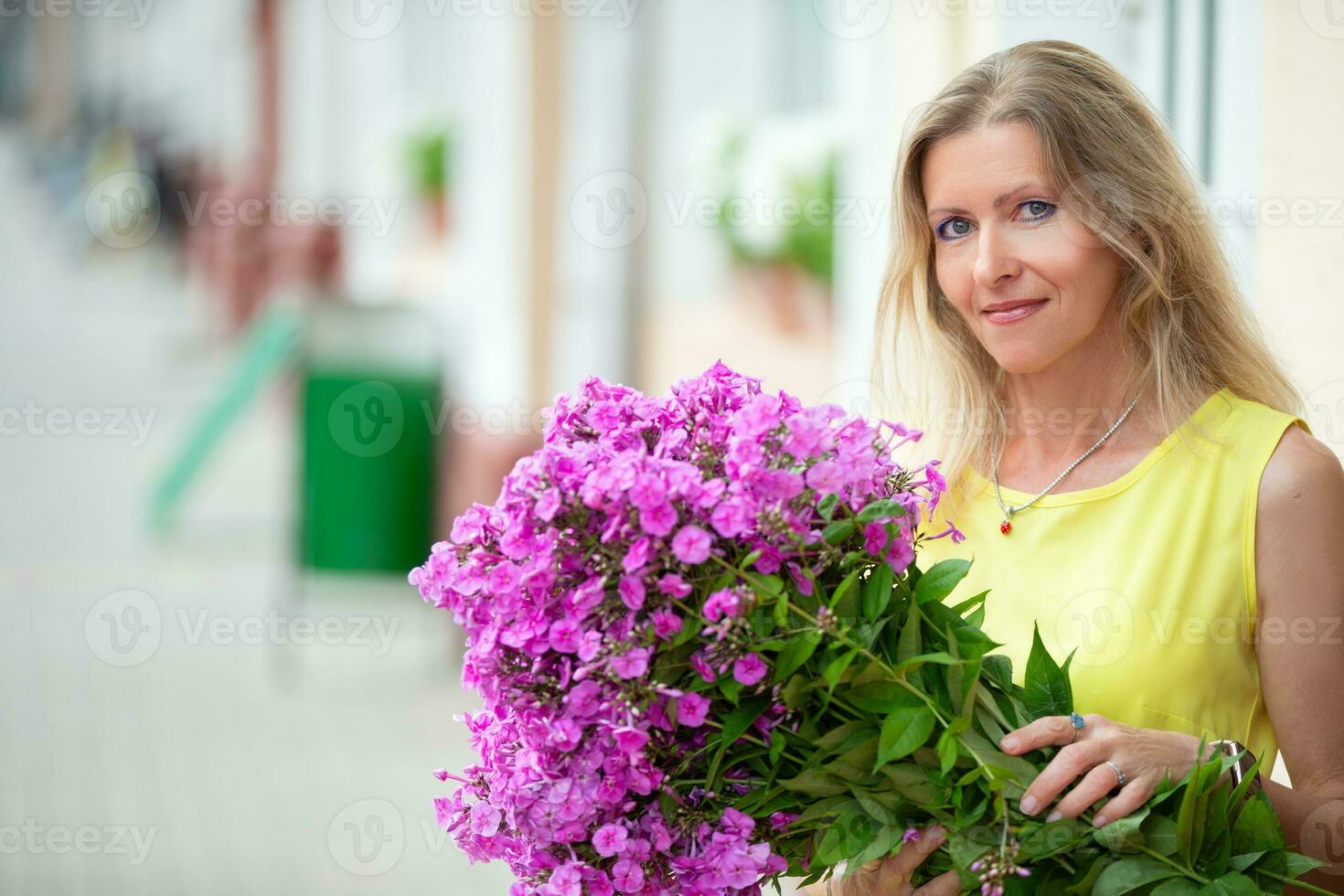 schön Alten Frau mit ein Strauß von Blumen. Alter Modell- blond mit Blau Augen lächelt und sieht aus beim das Kamera. ein glücklich Frau im ihr fünfziger Jahre. foto