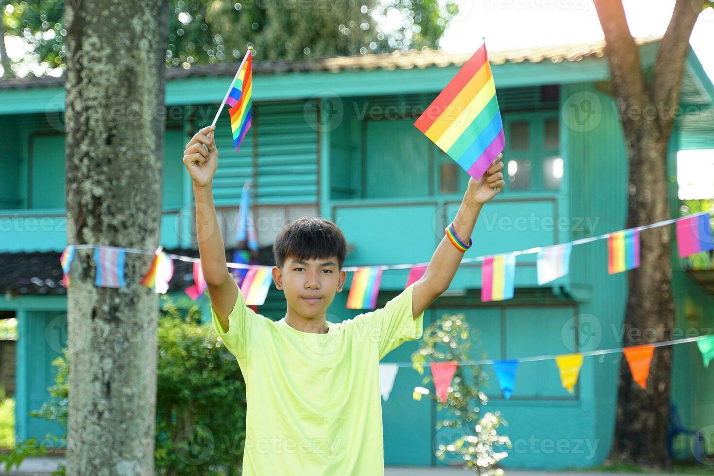 asiatisch Junge hält ein Regenbogen Flagge im Vorderseite von ein Haus dekoriert mit Regenbogen Flaggen während Stolz Monat zu Show lgbt Stolz und Identität. Sanft und selektiv Fokus. foto