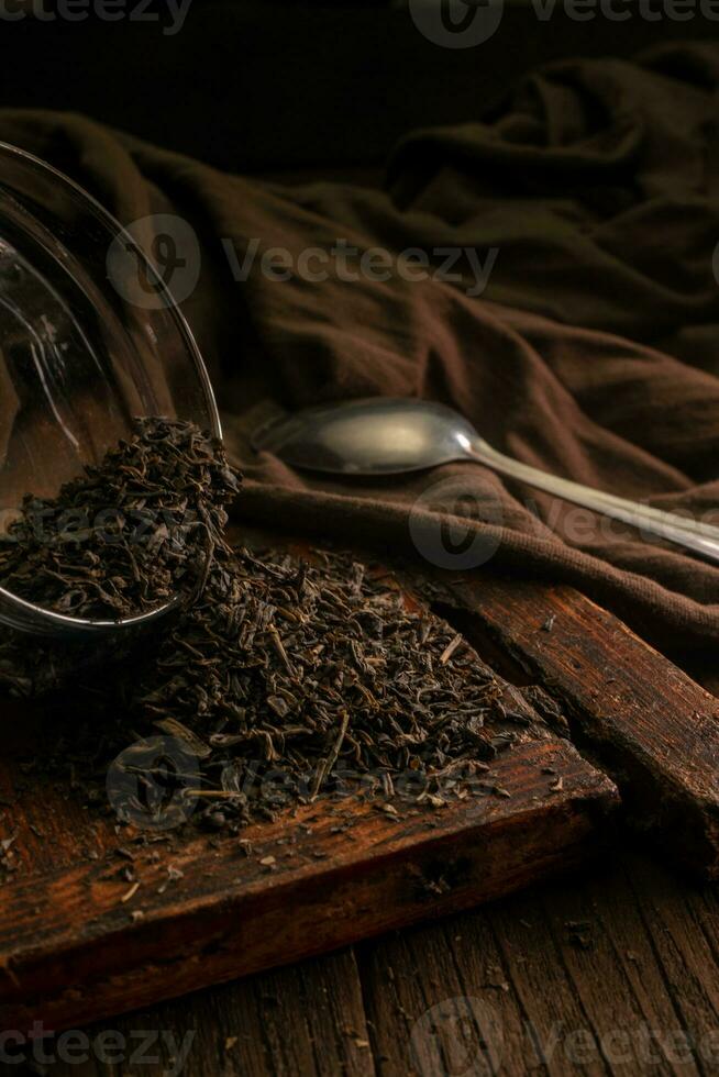 köstlich schwarz Blatt Tee ist verstreut auf ein hölzern braun Tabelle mit ein Löffel und ein Stoff auf ein dunkel Hintergrund foto