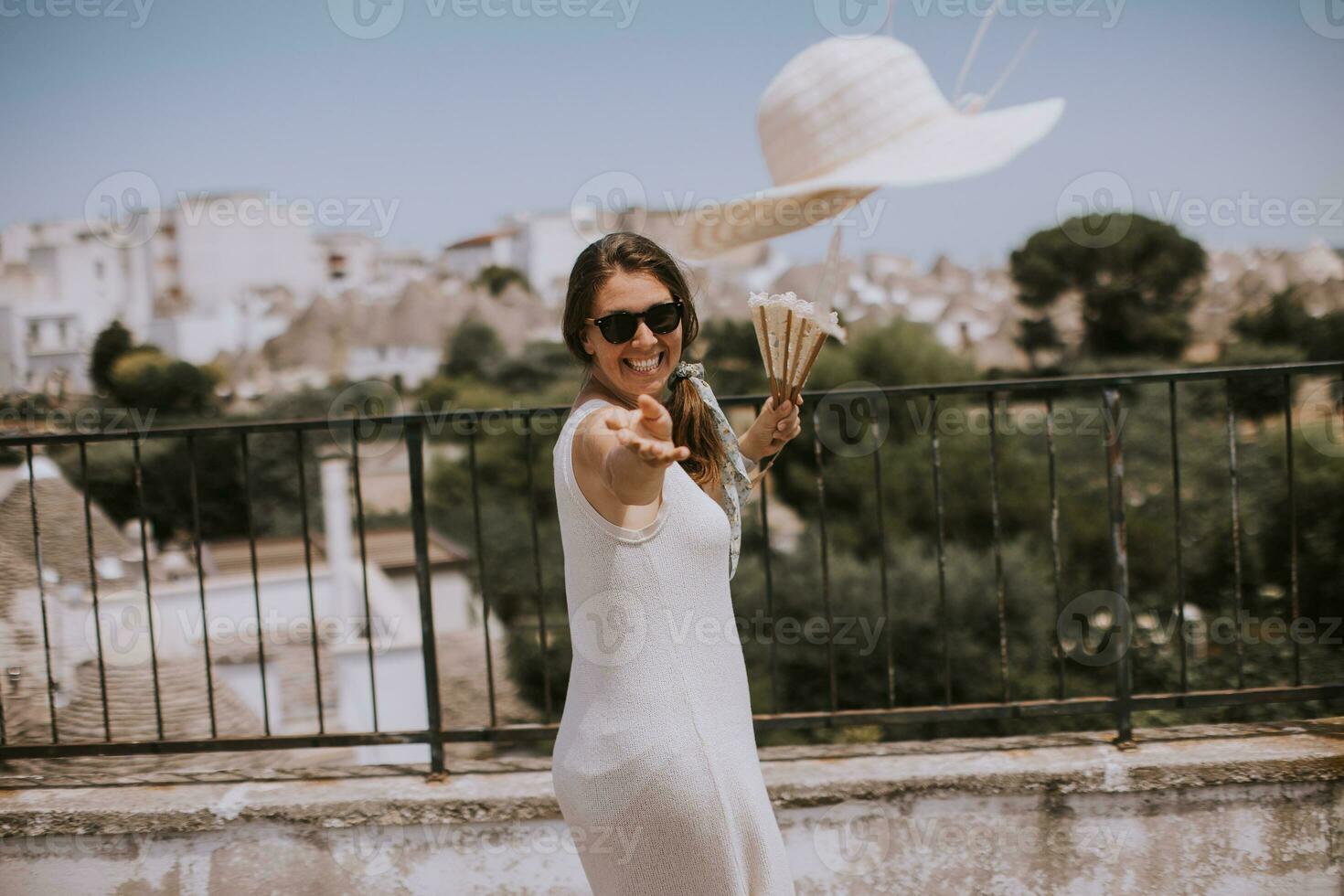 ein jung Frau im ein Weiß Kleid werfen Hut während Tourist Besuch im Alborebello, Italien foto