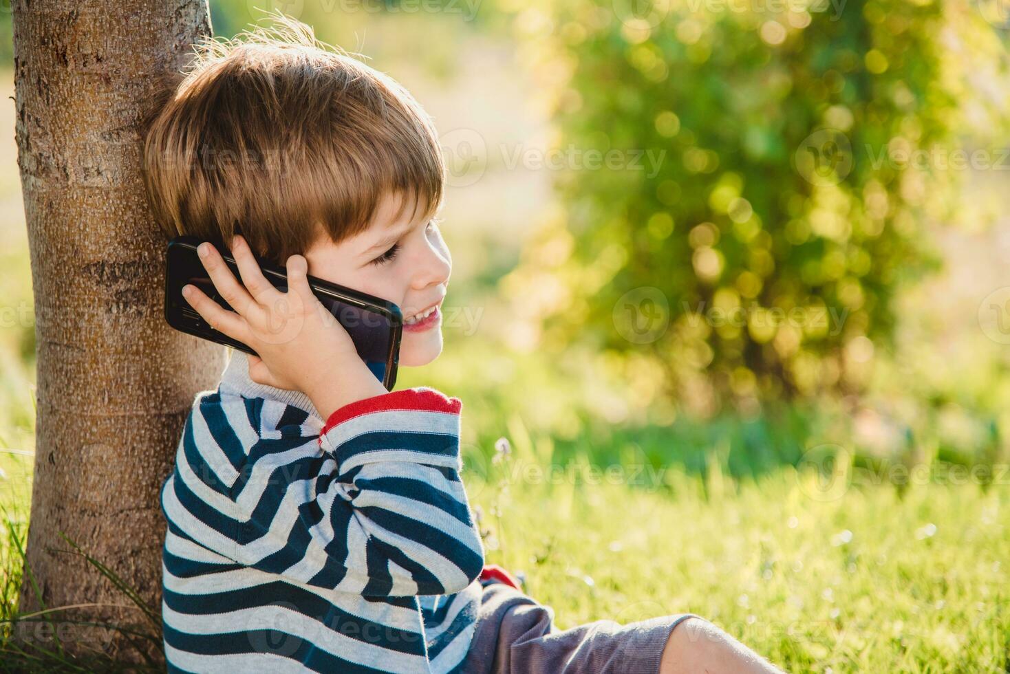 ein schön Kind Sitzung auf das Gras spricht durch Telefon im das Sommer- beim Sonnenuntergang. Junge kommuniziert auf Handy, Mobiltelefon foto