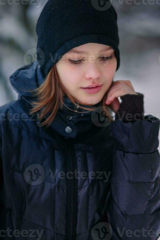 das Mädchen sieht aus Nieder mit ein Hand zu ihr Wange. herzlich gekleidet Kind im Winter auf das Straße. foto