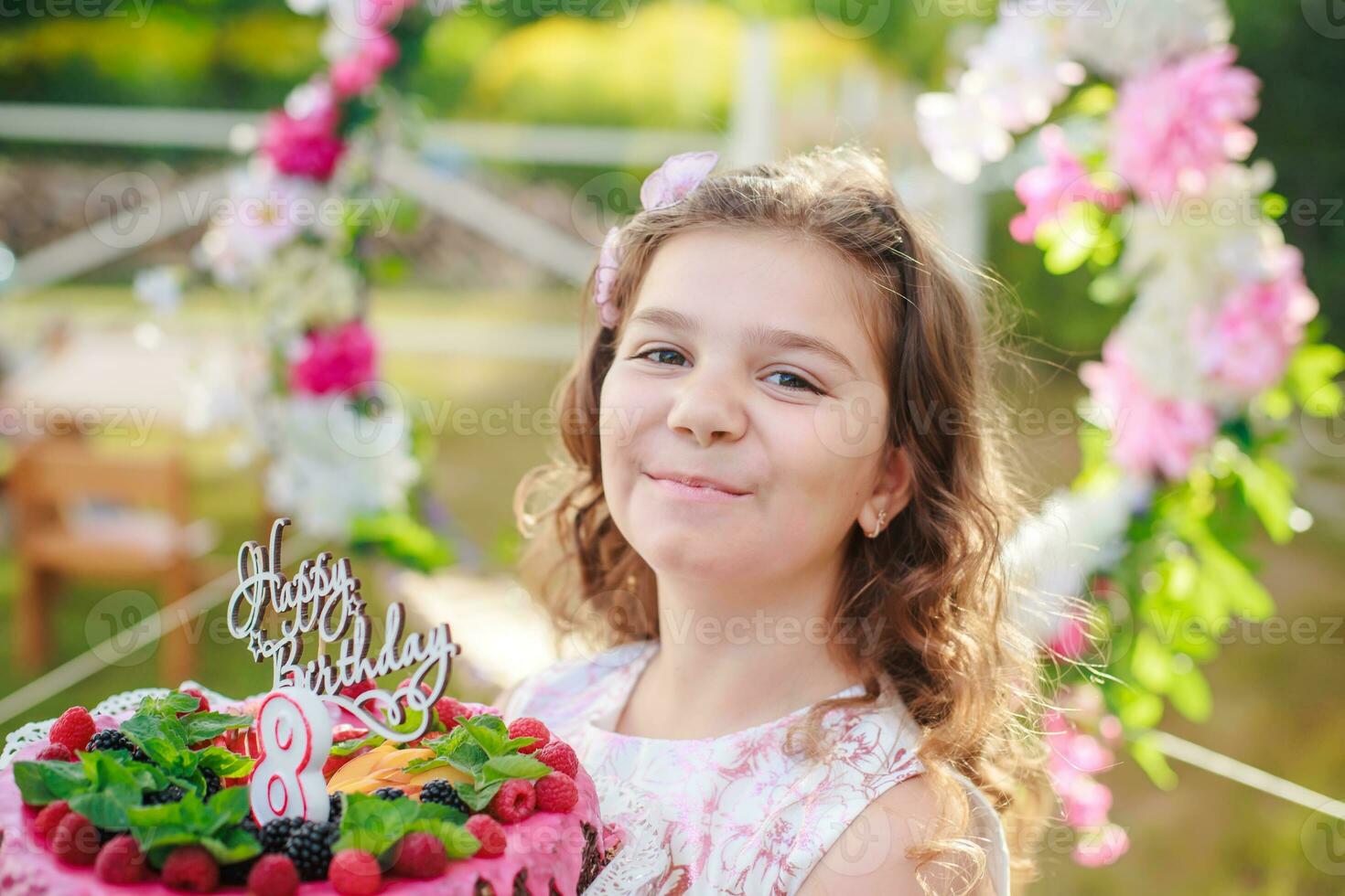 Mädchen halten Geburtstag Kuchen mit Nummer 8 und Inschrift glücklich Geburtstag. feiern zeremoniell Veranstaltungen auf Straße während Quarantäne. Kind im Hintergrund dekoriert Foto Zone von Blumen.