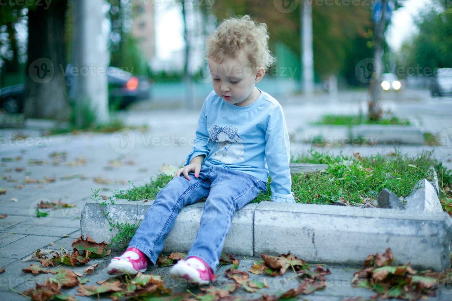 schön Mädchen mit lockig Haar Sitzung auf das Randstein im das Straße. Kind im Jeans und ein Blau Hemd spielen draußen foto