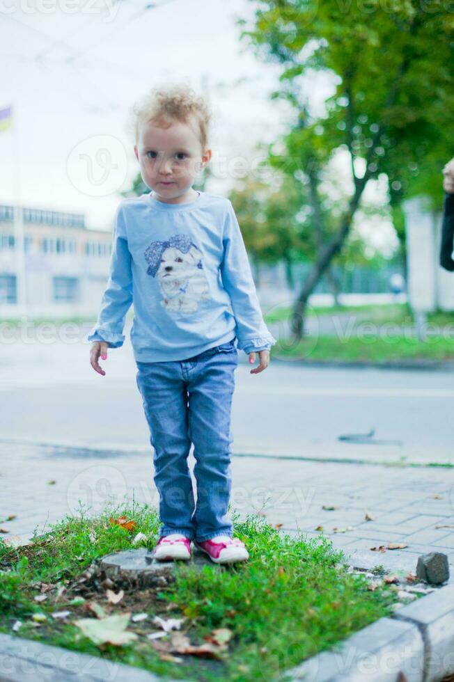 wenig schön Mädchen mit kurz lockig Haar Stehen auf ein Stumpf im das Stadt. Kind im Jeans und ein Blau Hemd spielen draußen foto