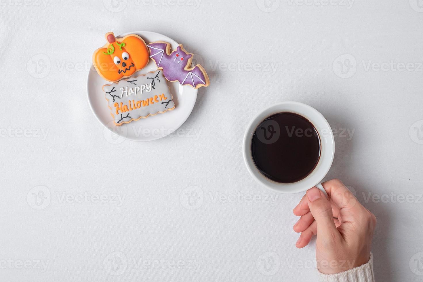 Frauenhand, die Kaffeetasse hält, während sie lustige Halloween-Plätzchen isst. Happy Halloween Day, Trick or Threat, Hallo Oktober, Herbstherbst, Traditions-, Party- und Urlaubskonzept foto