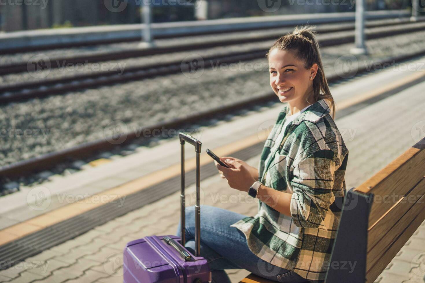 glücklich Frau mit Handy, Mobiltelefon Telefon während Sitzung auf ein Bank beim das Zug Bahnhof foto