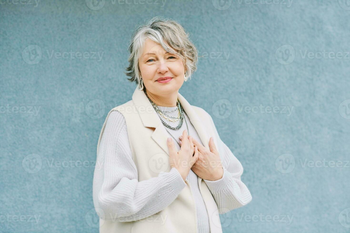 draussen Porträt von schön 55 - - 60 Jahr alt Frau posieren Nächster zu Blau Hintergrund Wand, halten Hände Nächster zu Brust, danken Sie Zeichen foto