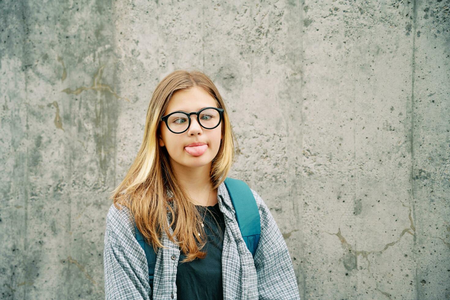 draussen Porträt von dumm jung Teenager Kind Mädchen ziehen ein Zunge, Kreuzung Augen Über ihr Nase, tragen Brille und Rucksack, posieren auf grau Mauer Hintergrund foto