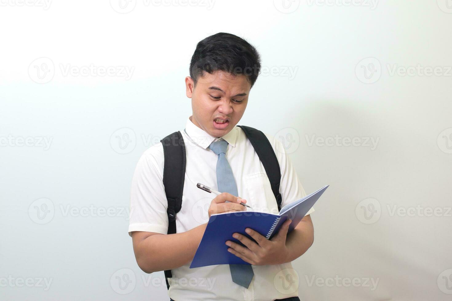 indonesisch Senior hoch Schule Schüler tragen Weiß Hemd Uniform mit grau Krawatte Schreiben auf Hinweis Buch mit Stift mit genervt und frustriert Ausdruck. isoliert Bild auf Weiß Hintergrund foto