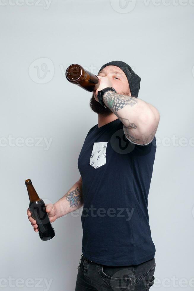 brutal bärtig männlich mit tätowiert Arm Getränke ein Bier von ein Flasche. foto