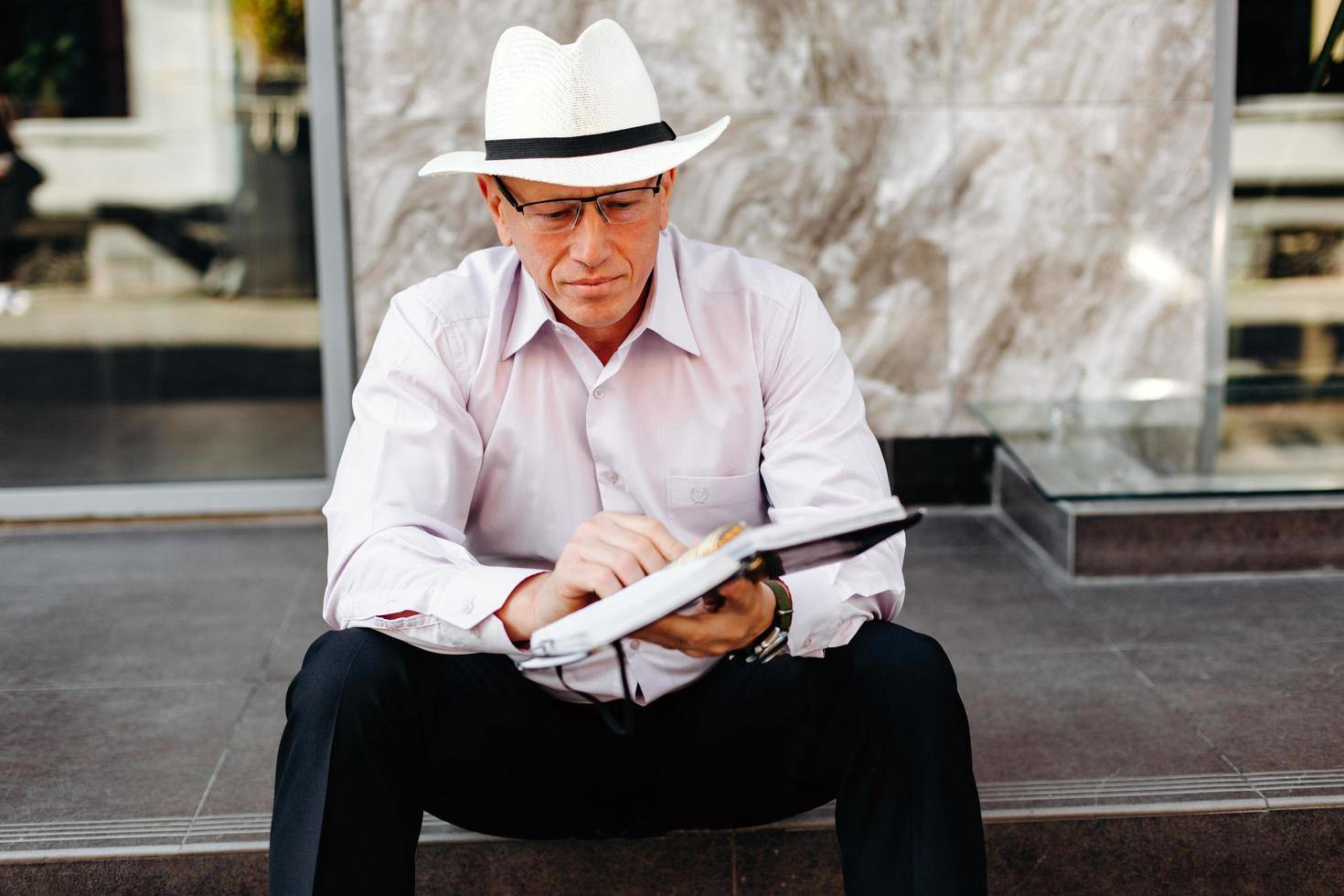 älterer Mann mit Hut, der auf dem Bürgersteig sitzt und ein Notizbuch in den Händen hält - Bild foto
