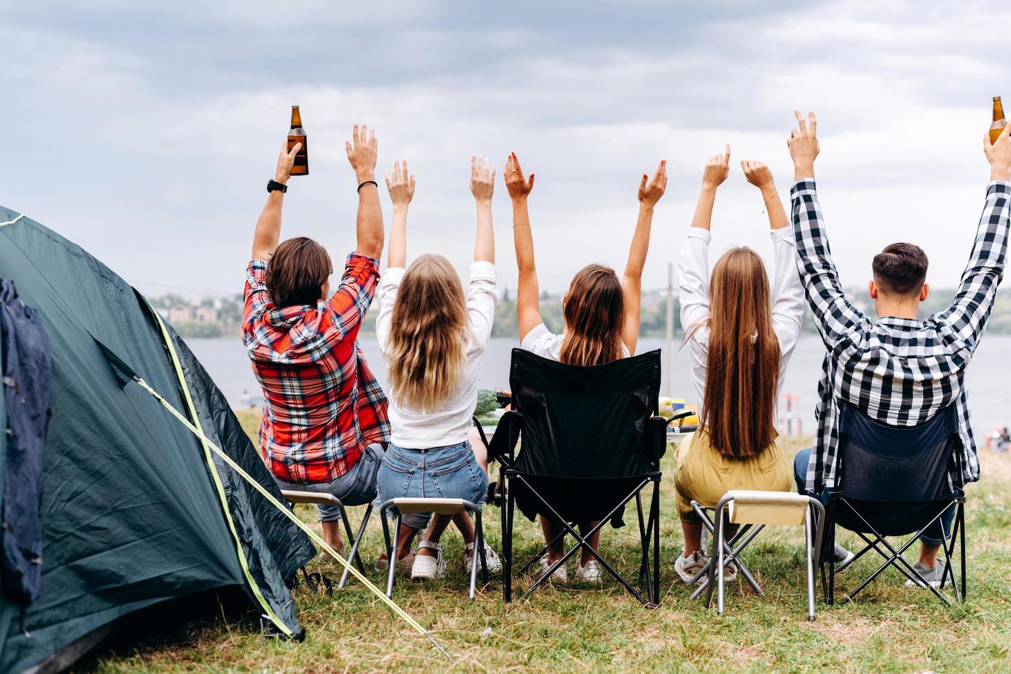 eine Gesellschaft von Freunden isst auf dem Campingplatz zu Mittag. sie hände hoch. - Rückansicht foto