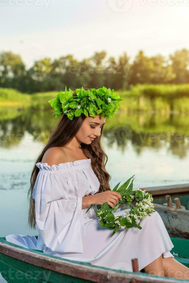 das Nymphe mit lange dunkel Haar im ein Weiß Jahrgang Kleid Sitzung im ein Boot im das Mitte von das Fluss. foto