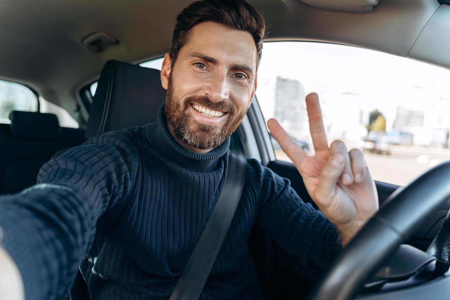 Selfie machen. Gut aussehender bärtiger Mann, der Kamera hält und gestikuliert, während er im Auto am Fahrersitz sitzt und mit Freude ein Selbstporträt macht foto