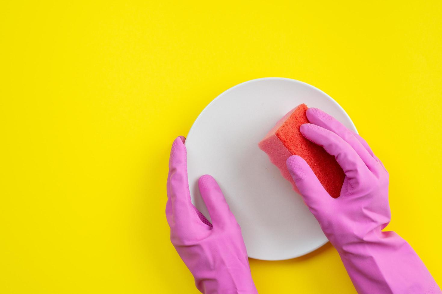 Nahaufnahmehände tragen in Latexhandschuhen, die einen Küchenschwamm und einen Teller halten. Draufsicht auf gelbem Hintergrund beim Geschirrspülen foto