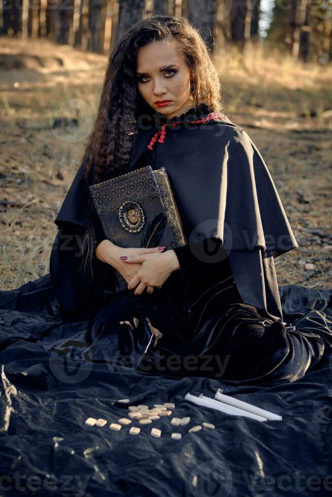 Hexe im schwarz Kleid mit Kap und Haube. posieren im Kiefer Wald. Sitzung auf dunkel Decke mit Bücher, Kerzen und Runen auf Es. voll Länge. foto