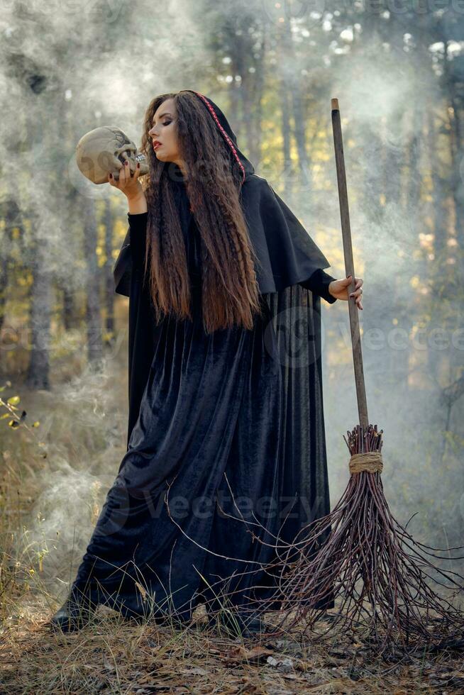 Hexe im Schwarz, lange Kleid mit Kap und Haube. posieren im Rauch Kiefer Wald. halten ein Schädel und ein Besen. Zauber, Magie und Hexerei. voll Länge. foto