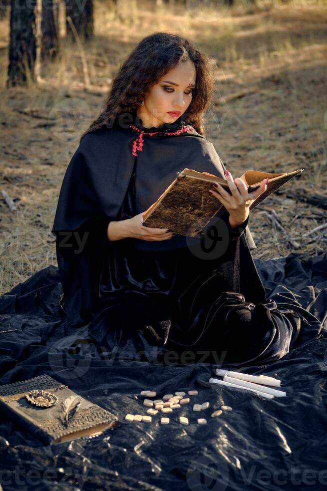 Hexe im schwarz Kleid mit Kap und Haube. posieren im Kiefer Wald. Sitzung auf dunkel Decke mit Bücher, Kerzen und Runen auf Es. voll Länge. foto