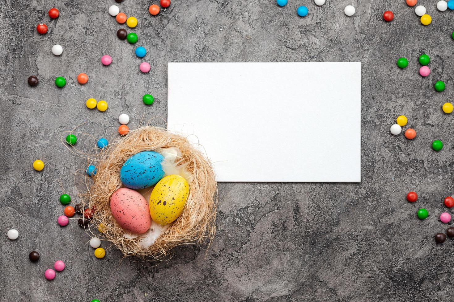 Auf grauem Gipsgrund liegt ein Vogelnest mit bunten Eiern, einer Blanko-Osterkarte und Süßigkeiten foto