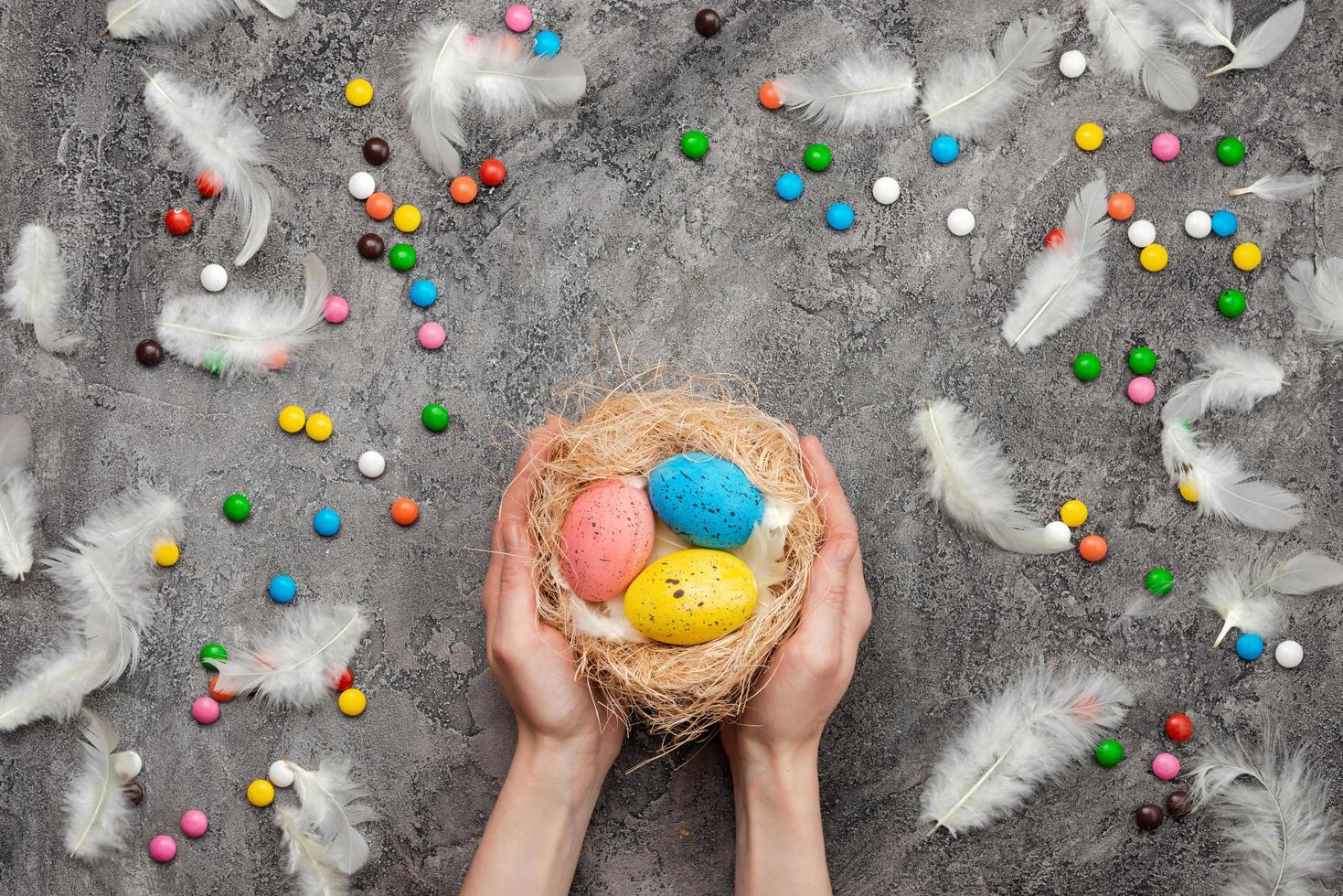 Osterhintergrund, weibliche Hände, die ein Nest mit bunten Eiern halten. Süßigkeiten und Federn verstreut foto