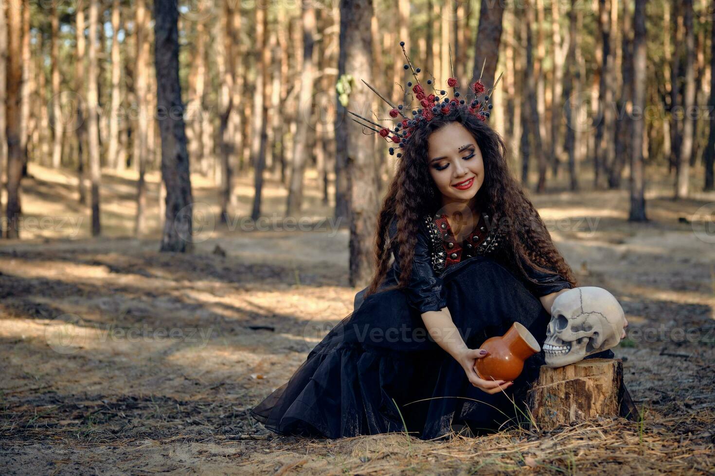 Hexe im Schwarz, lange Kleid, mit rot Krone im Haar. geben Wasser zu Schädel von Topf während Sitzung im Kiefer Wald. Zauber, Hexerei. voll Länge. foto