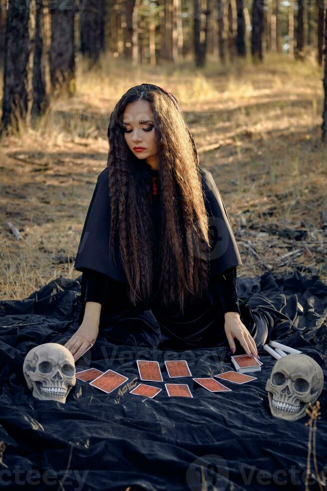 Hexe im schwarz Kleid mit Kap und Haube. posieren im Kiefer Wald. Sitzung auf dunkel Decke mit Wahrsagen Karten und Schädel auf Es. voll Länge. foto