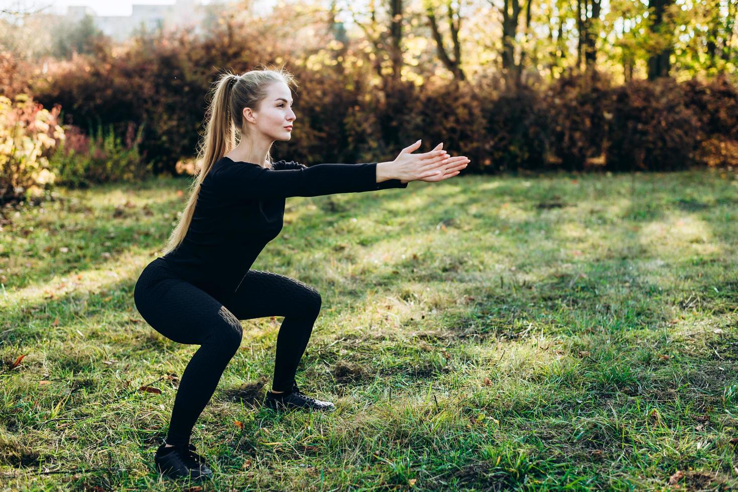 Schönes Mädchen in schwarzer Sportbekleidung führt Kniebeugen im Park durch, Seitenansicht. foto