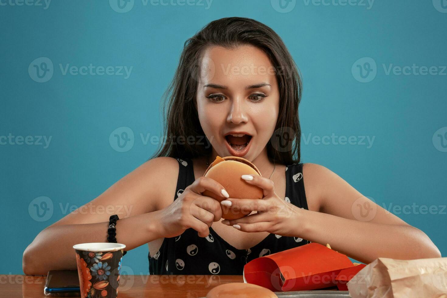 Nahansicht Porträt von ein Frau im ein schwarz Kleid posieren Sitzung beim das Tabelle mit Burger, Französisch Fritten und trinken. Blau Hintergrund. schnell Lebensmittel. foto