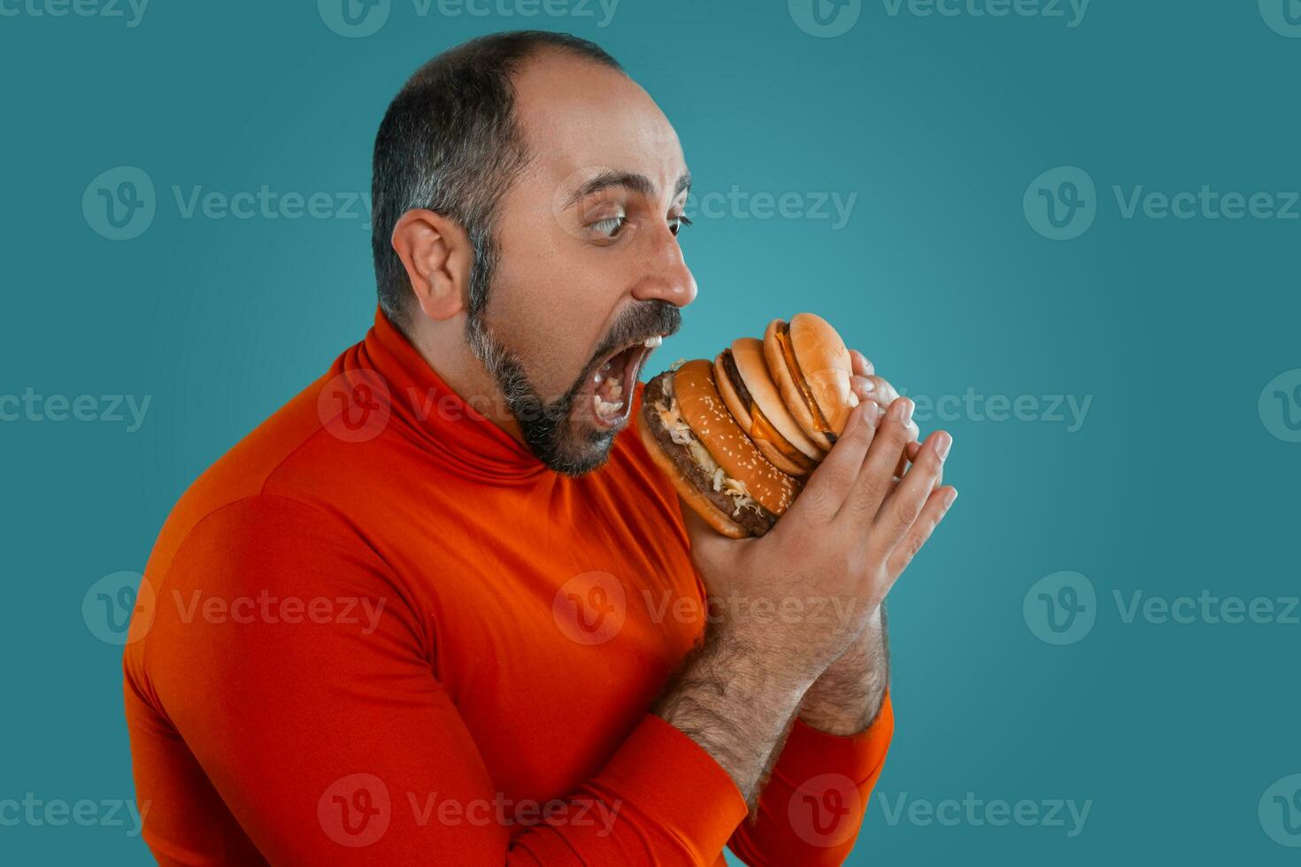 Nahansicht Porträt von ein mittleren Alters Mann mit Bart, gekleidet im ein rot Rollkragen, posieren mit Burger gegen ein Blau Hintergrund. schnell Lebensmittel. foto