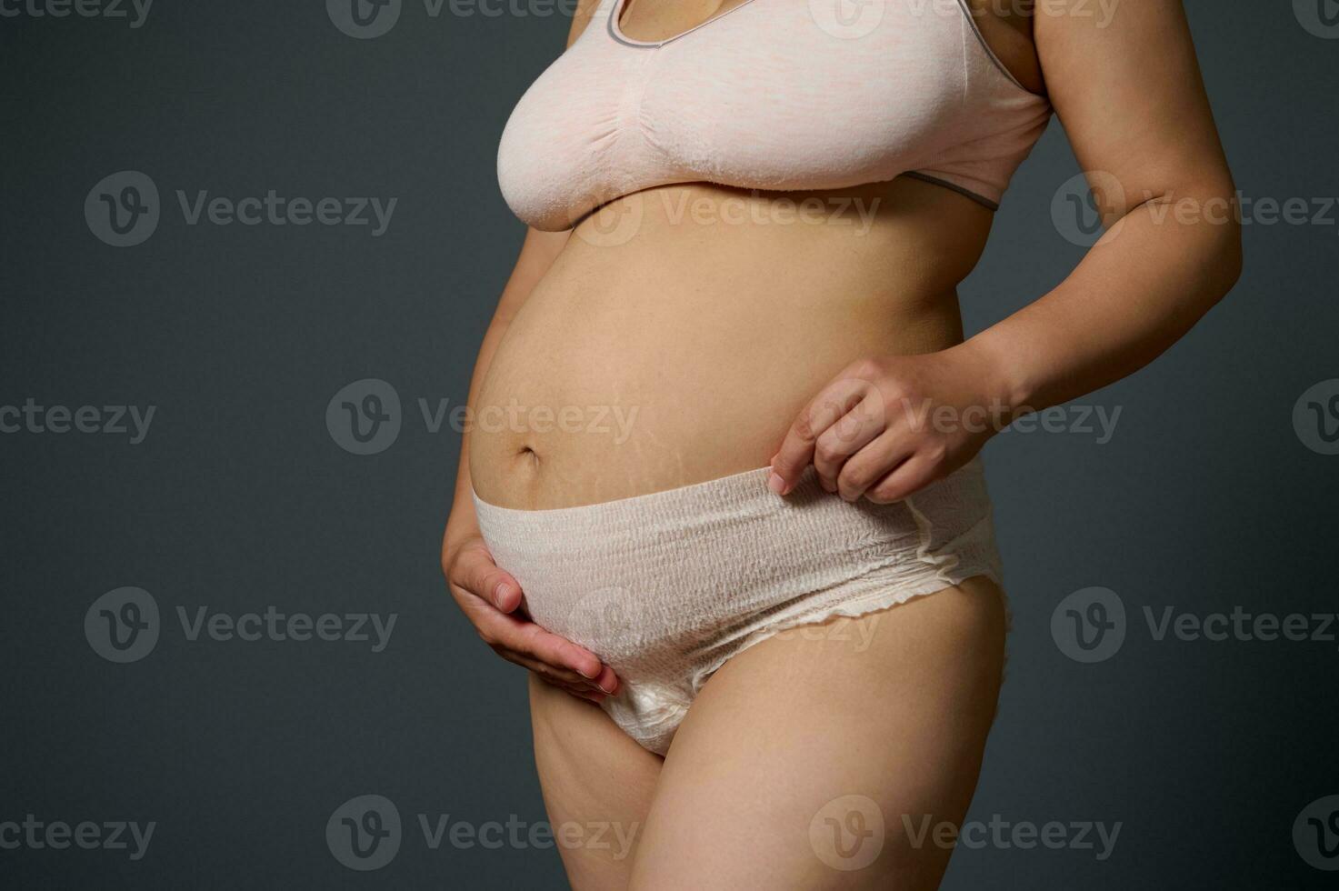 Nahansicht Mutter halten ihr postnatal Bauch, Stehen isoliert auf grau Studio Hintergrund, gekleidet im Mutterschaft Unterwäsche foto