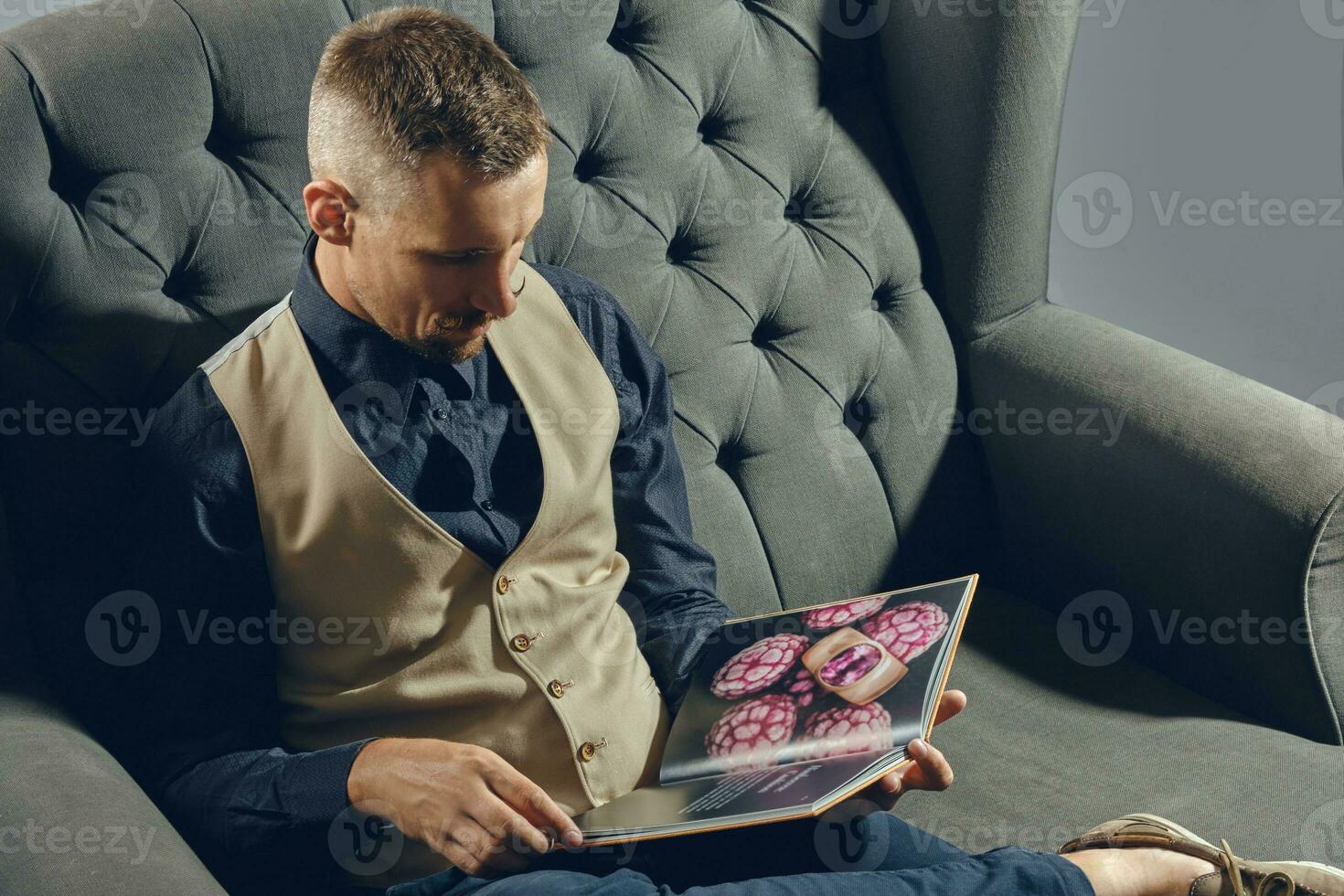 Mann mit stilvoll Schnurrbart, gekleidet im schwarz Hemd und Hose, Beige Weste ist Sitzung auf dunkel Sofa, lesen ein Zeitschrift. grau Hintergrund, Nahansicht. foto