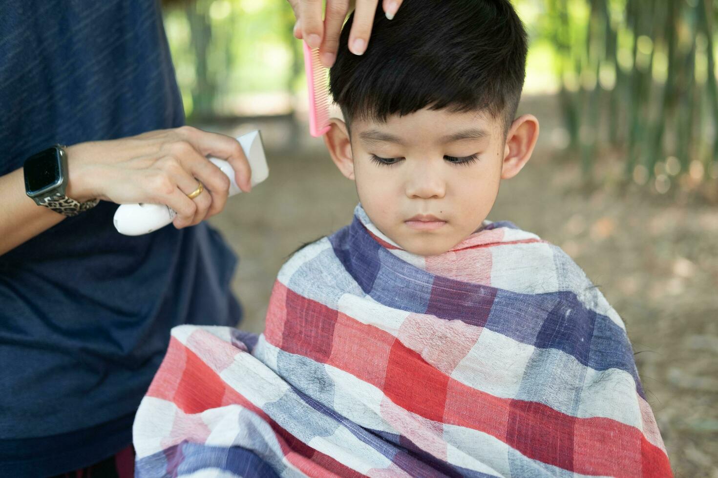 Barbier Schneiden Haar von ein asiatisch Junge im ein öffnen Raum gefüllt mit Bäume. foto