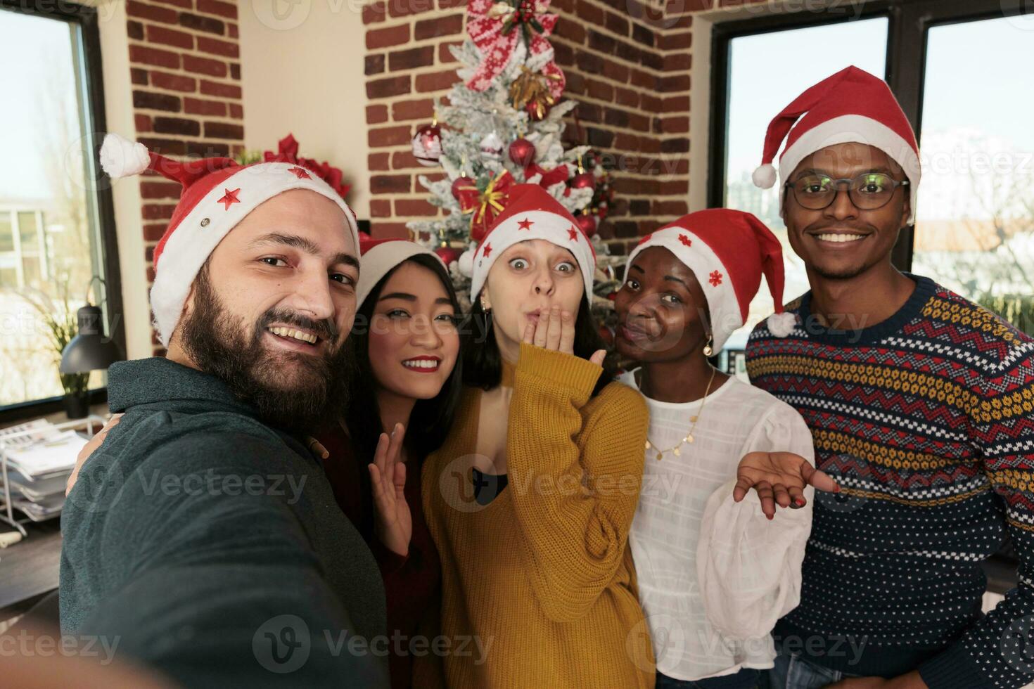 lächelnd vielfältig Männer und Frauen Kollegen tragen Santa Hüte posieren zum Selfie zusammen im Büro mit Weihnachten Dekorationen. heiter Mitarbeiter nehmen Gruppe Foto beim Neu Jahr korporativ Party