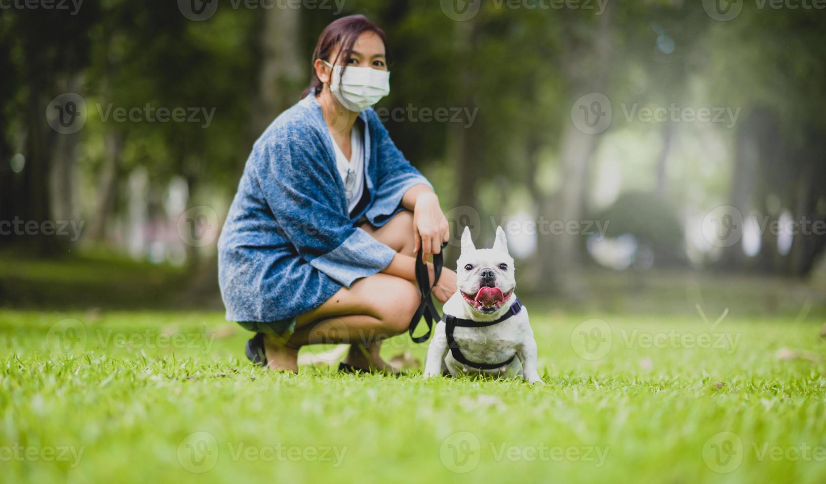 eine frau, die eine medizinische maske trägt, die mit einer weißen französischen bulldogge auf dem rasen sitzt. foto