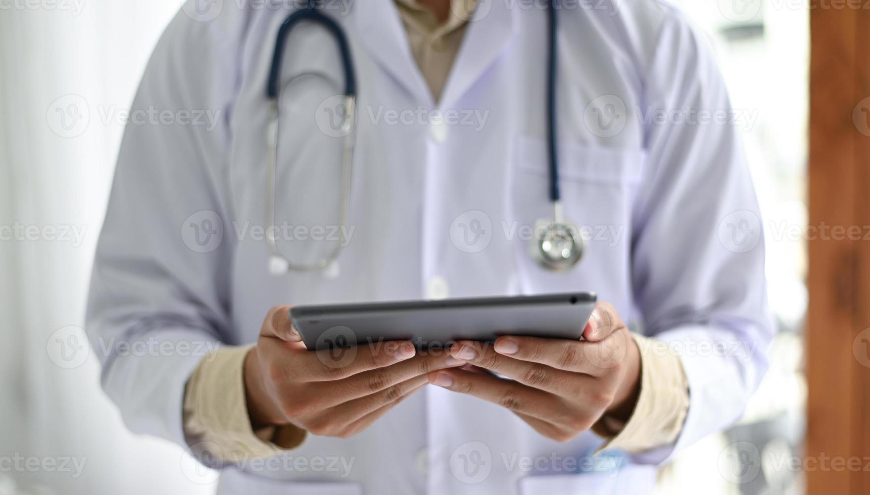 Ausgeschnittene Aufnahme eines Mediziners in einem Laborkittel mit einem Stethoskop, das eine Tablette in der Hand hält. foto