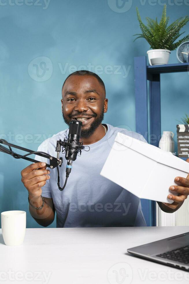 lächelnd Blogger Aufzeichnung hergeben Video und Werbung Produkt online Porträt. afrikanisch amerikanisch vlogger halten Box mit Geschenk zum Abonnenten, zeigen Wettbewerb Preis- und suchen beim Kamera foto