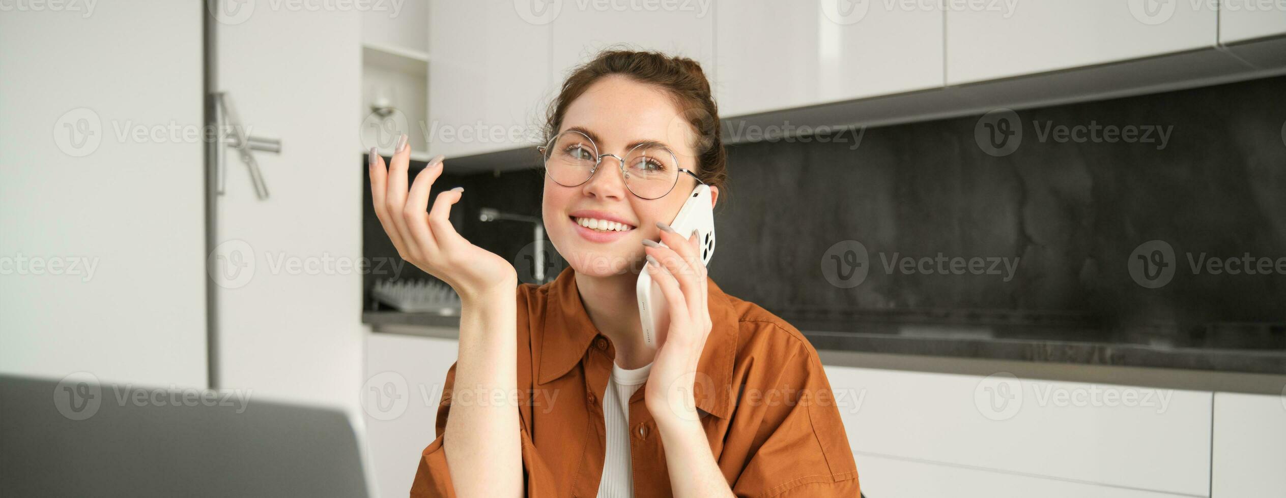 Porträt von jung Frau, Geschäft Inhaber Arbeiten von heim, Schüler Herstellung ein Telefon Forderung, Sitzung im Küche mit Laptop, reden zu jemand foto