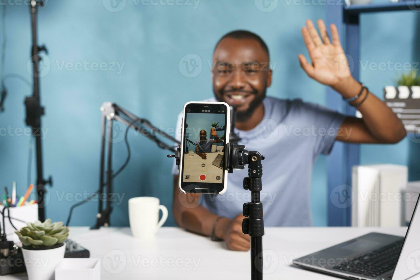 lächelnd Streamer Rundfunk- Leben mit Smartphone auf Stativ und Fachmann Ausrüstung. afrikanisch amerikanisch Blogger Gruß Internet Kanal Publikum und Aufzeichnung Video auf Handy, Mobiltelefon Telefon schließen oben foto