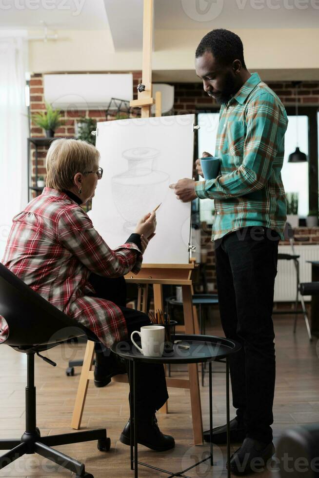 afrikanisch amerikanisch Kerl Kunst Lehrer Lehren Senior Frau zu zeichnen im Klasse, Stehen in der Nähe von Staffelei und erklären Skizzierung Techniken. Lehrer assistieren reifen Schüler während Zeichnung Lektion foto