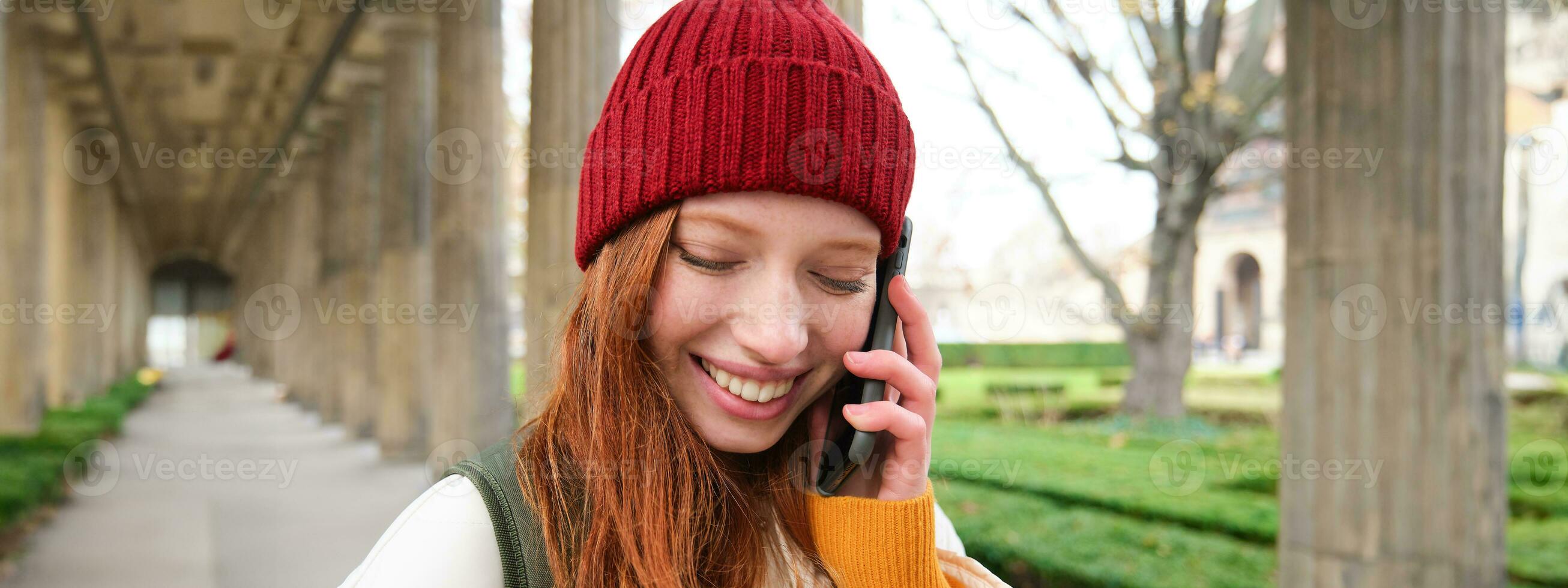 Porträt von Rothaarige europäisch Mädchen im rot Hut, macht ein Telefon Forderung, Spaziergänge im Stadt und Gespräche zu Freund auf Smartphone foto