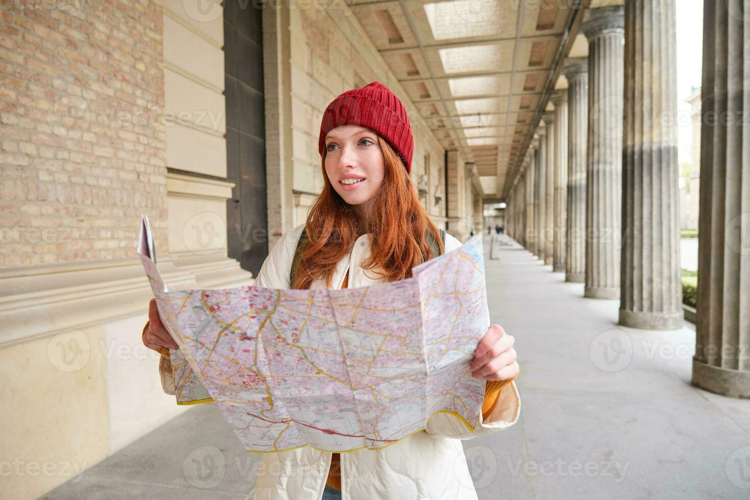lächelnd jung Rothaarige Frau im rot Hut, sieht aus beim Papier Karte zu aussehen zum Tourist Attraktion. Tourismus und Menschen Konzept. Mädchen erforscht Stadt, versucht zu finden Weg foto