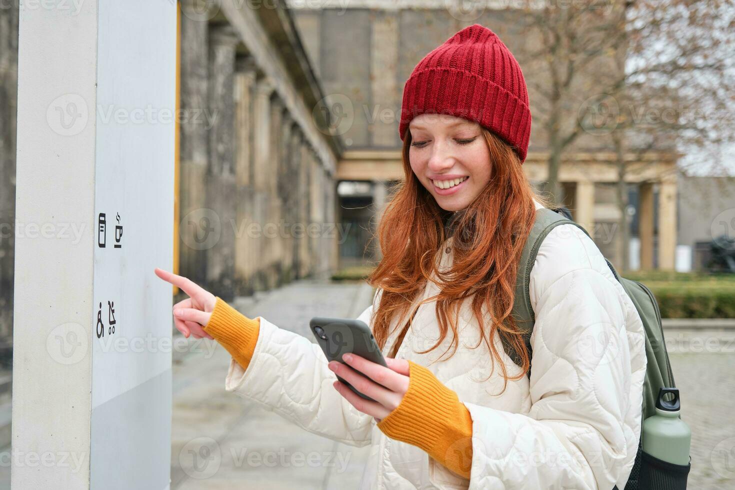 lächelnd Rothaarige Mädchen Tourist, Spaziergänge um Stadt und erforscht Beliebt Sehenswürdigkeiten, Besichtigung, halten Smartphone, suchen beim ihr Handy, Mobiltelefon App und prüft mit Stadt Karte foto