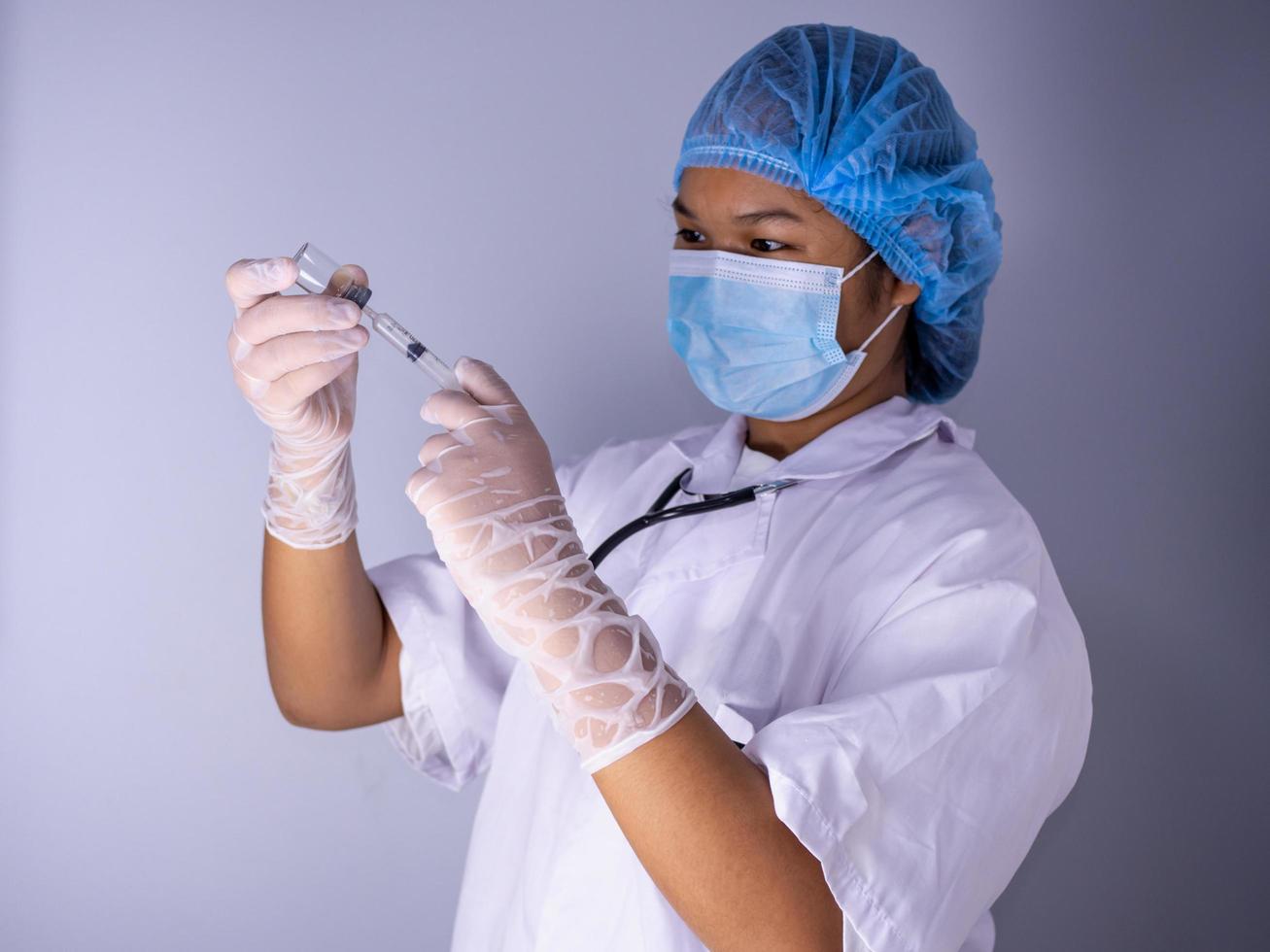 Studioporträt einer Ärztin, die eine Maske trägt und einen Hut trägt. In der Hand hielt er eine Flasche Impfstoff und eine Schlinge Spritzen. stehend auf weißem Hintergrund. Studioaufnahmehintergrund, Covid-19-Konzept foto