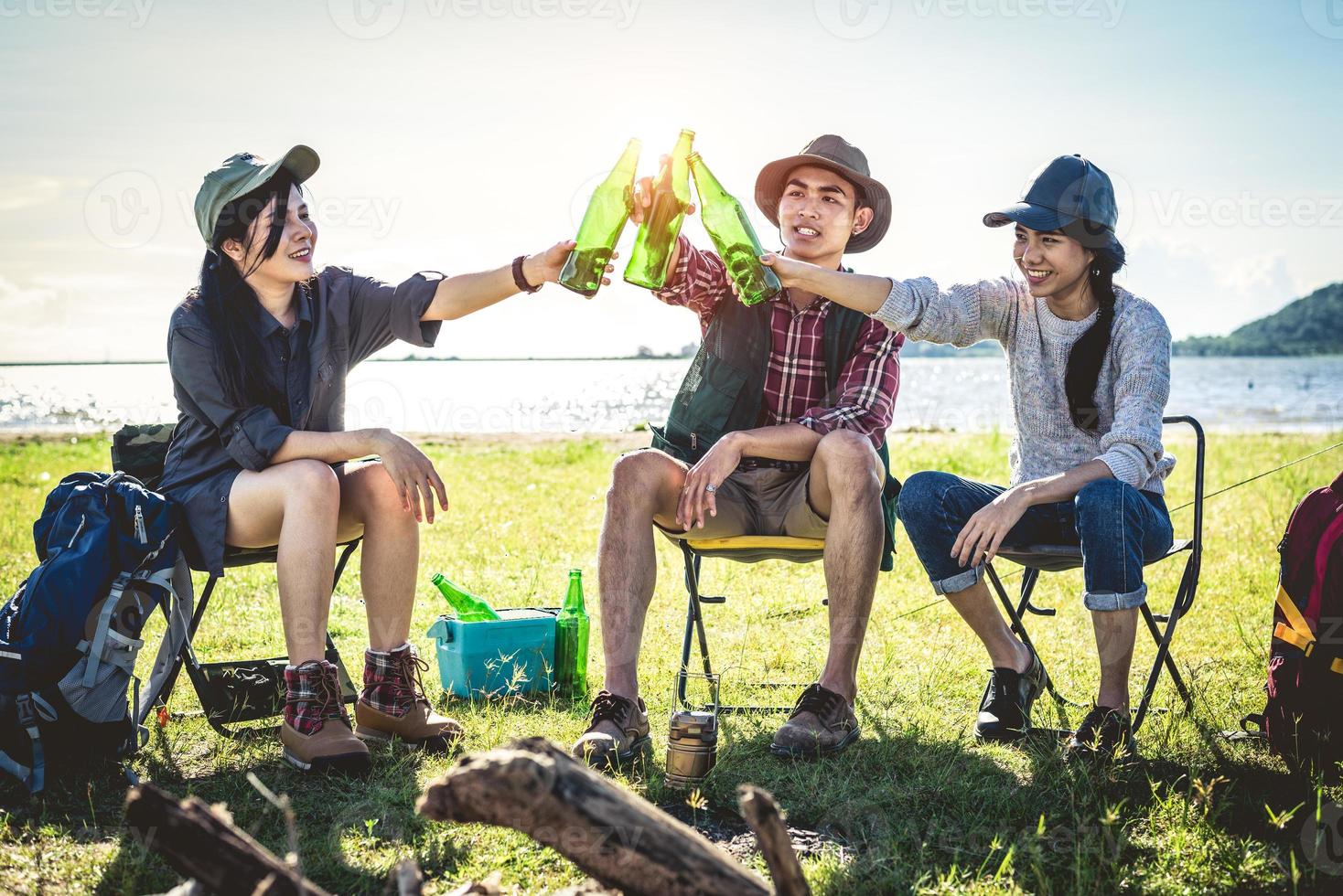 Gruppe junger asiatischer Freunde genießen Picknick und Party am See mit Campingrucksack und Stuhl. junge Leute, die Bierflaschen anstoßen und jubeln. Menschen und Lebensstile Konzept. Hintergrundthema im Freien foto