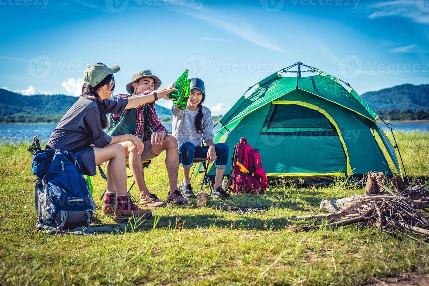 Gruppe junger asiatischer Freunde genießen Picknick und Party am See mit Campingrucksack und Stuhl. junge Leute, die Bierflaschen anstoßen und jubeln. Menschen und Lebensstile Konzept. Hintergrundthema im Freien foto