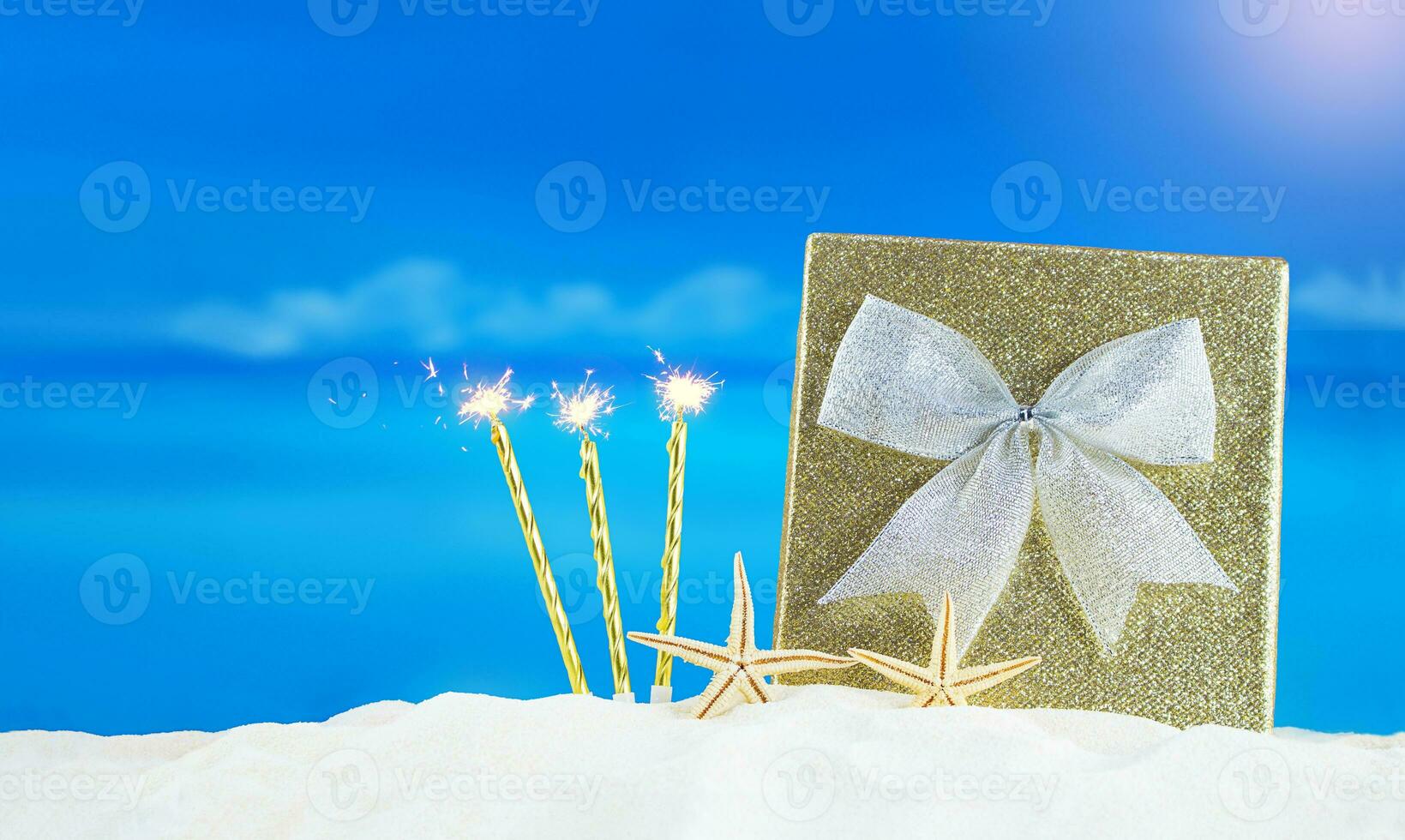 golden glänzend Box mit Silber Bogen, Kerzen mit Flammen und Seestern auf Sand von Strand. Weihnachten, Neu Jahr, Geburtstag, International Damen und Mutter Tag, Urlaub. im heiß Länder. Kopieren Raum foto