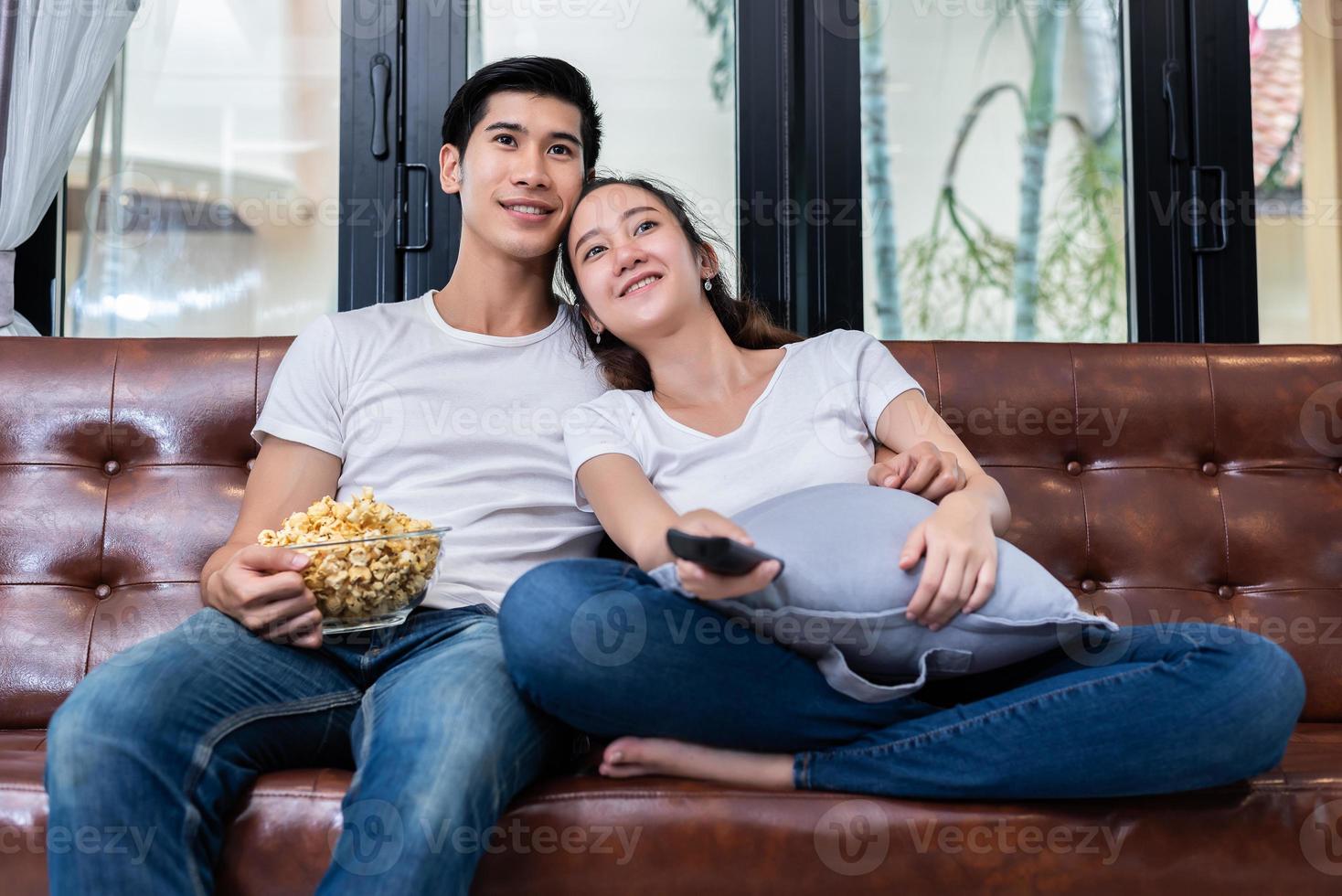 asiatische Paare, die fernsehen und zusammen Popcorn auf dem Sofa in ihrem Haus essen. Menschen und Lebensstile Konzept. Happy Home und Aktivitätsthema foto