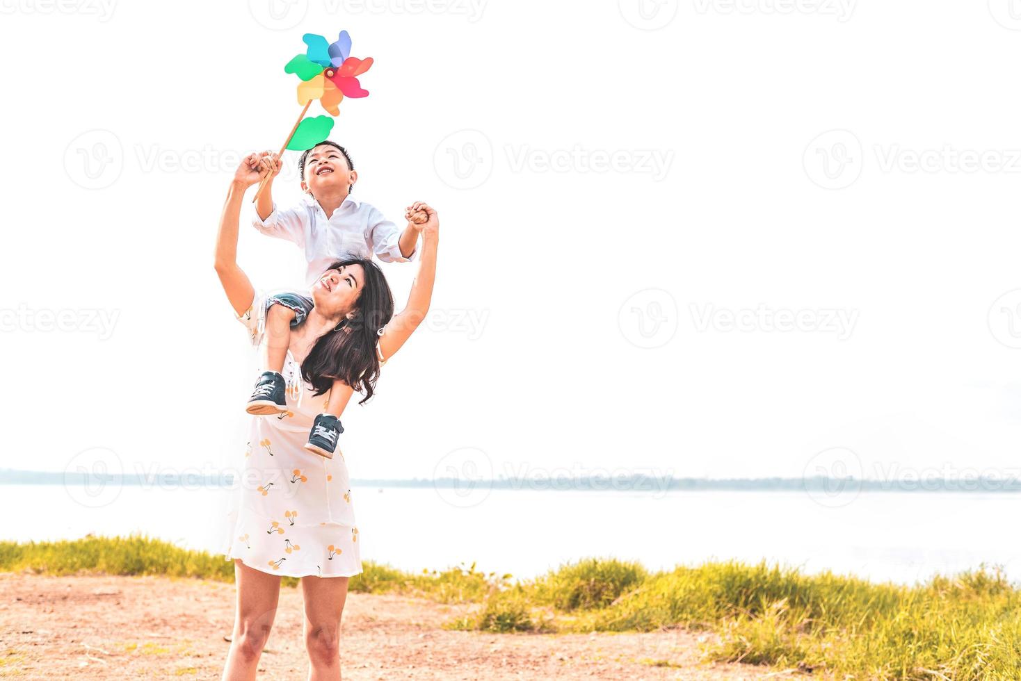 Kleiner asiatischer Junge, der auf seiner Superpower-Mutter auf der Wiese in der Nähe des Sees zurückreitet. Mutter und Sohn spielen zusammen. am Muttertag feiern und das Konzept schätzen. Sommer-Menschen und Lifestyle-Thema foto