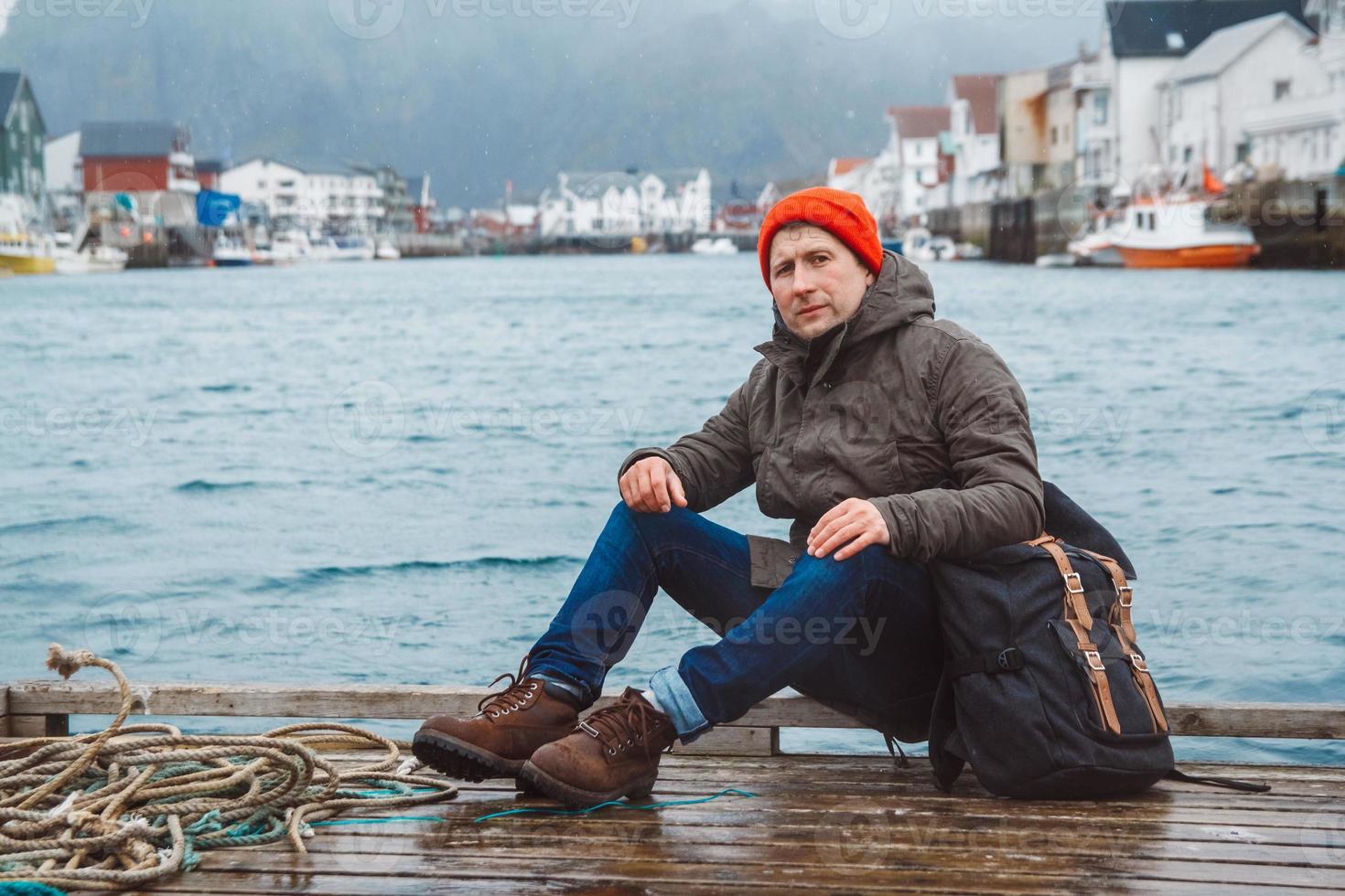 Reisender Mann mit Rucksack sitzt auf Woode Pier auf dem Hintergrund der Fischerhäuser Berg und See foto